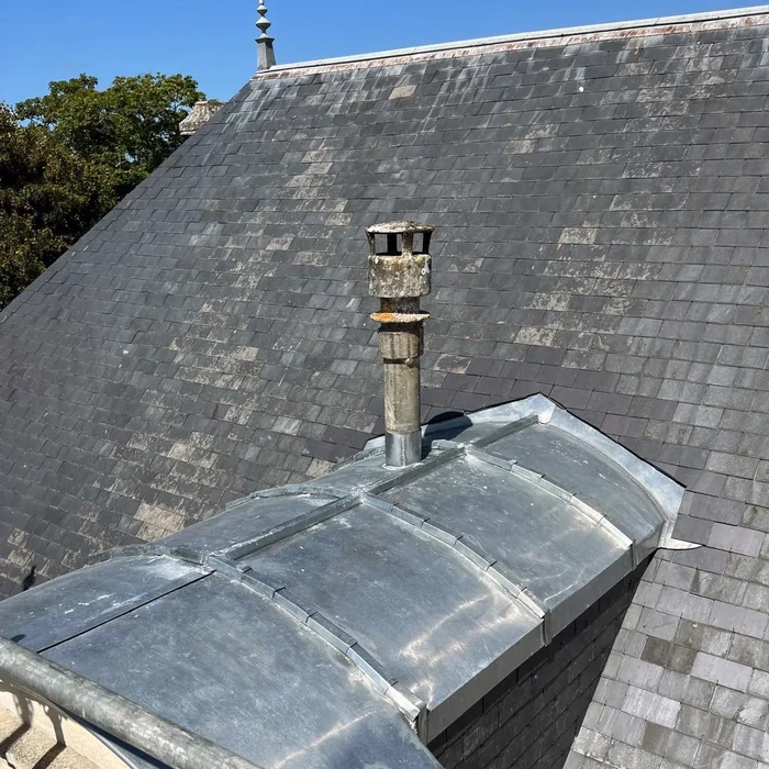 Vue aérienne d'une toiture en ardoises avec cheminée en pierre et souche cylindrique présentant des traces d'usure et mousses